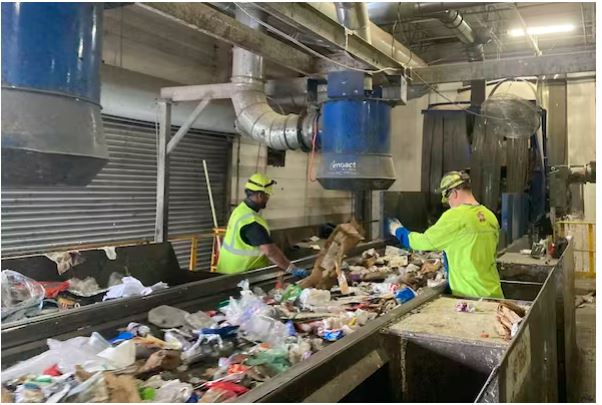 دو کارگر با رنگ زرد روشن در کنار یک تسمه نقاله پوشیده از پلاستیک در یک مرکز بازیافت ایستاده اند.